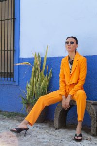 Fashionable woman in an orange suit sitting outside on a bench against a colorful wall.