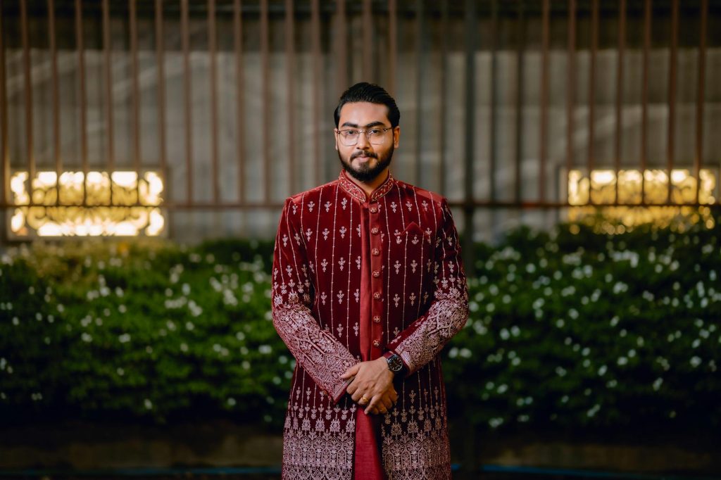 Man wearing a maroon sherwani stands confidently outdoors at night.