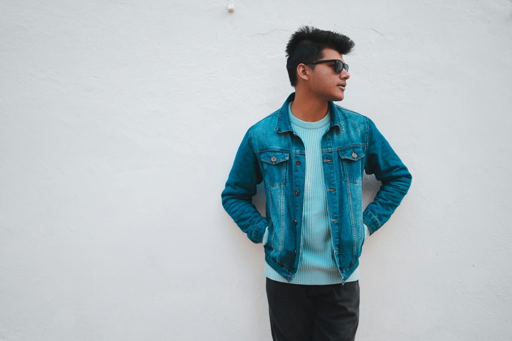 Asian male model wearing a denim jacket posing against a white wall, exhibiting cool urban style.