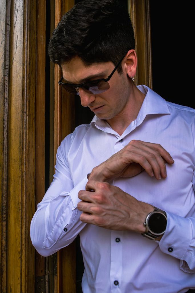 Man in white shirt adjusting cufflinks while standing near a wooden door, exuding style and elegance.